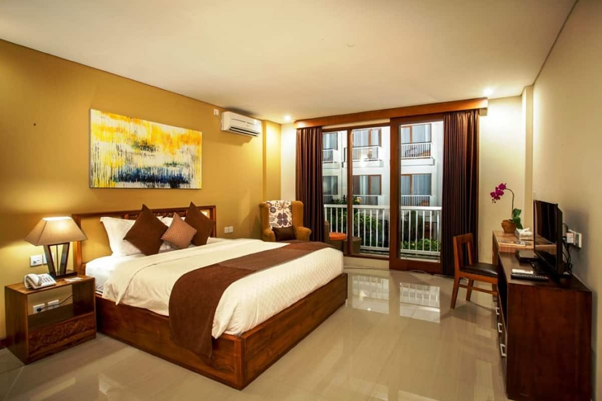 Ein Hotelzimmer mit einem großen Bett, einem Schreibtisch mit Computer, einem Sessel, einem Fernseher und einem Balkon mit Blick auf Gebäude.