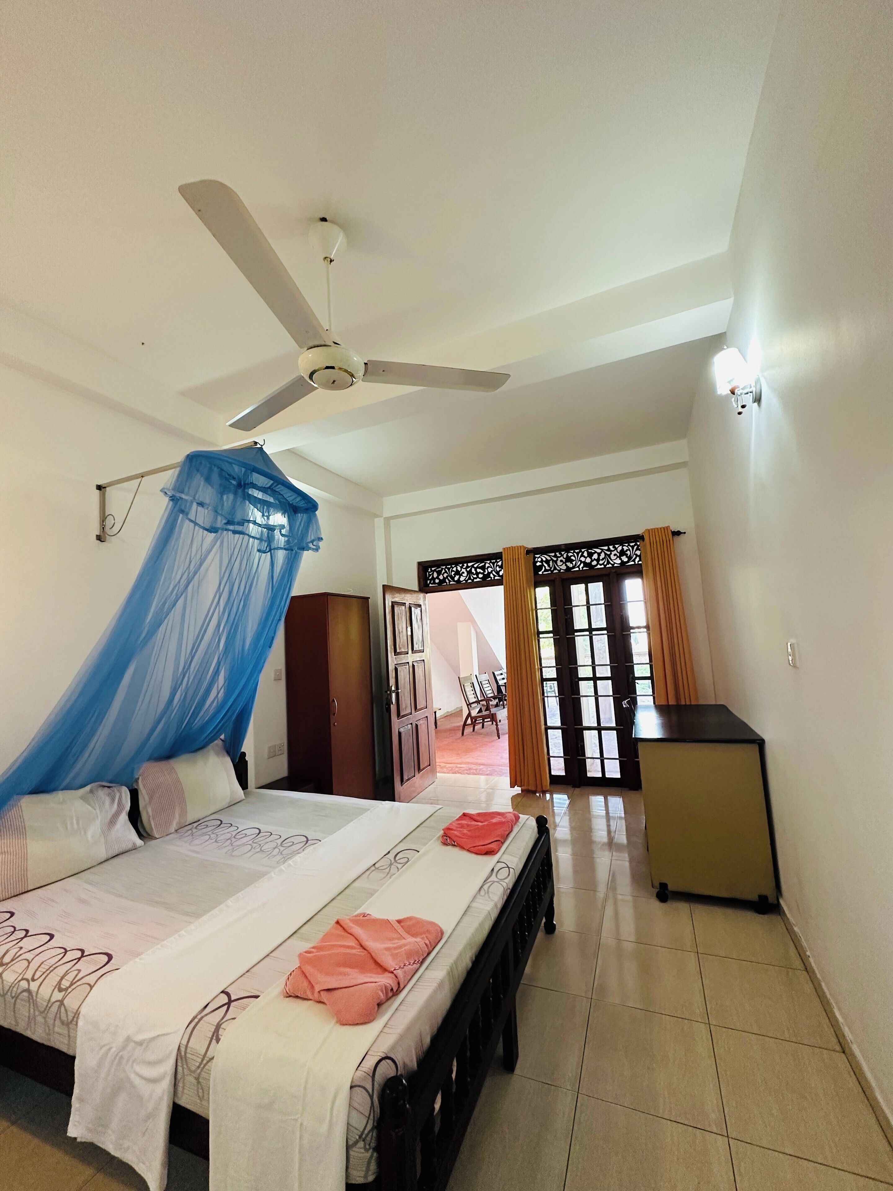 Une chambre à coucher avec un lit, un ventilateur de plafond, un bureau et une vue sur l’extérieur, visible à travers une porte.