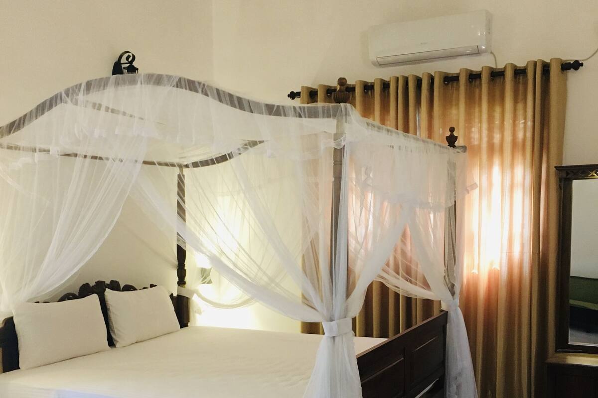A bed with a canopy, a bedside table, a chair, a red rug, and an air conditioning unit.