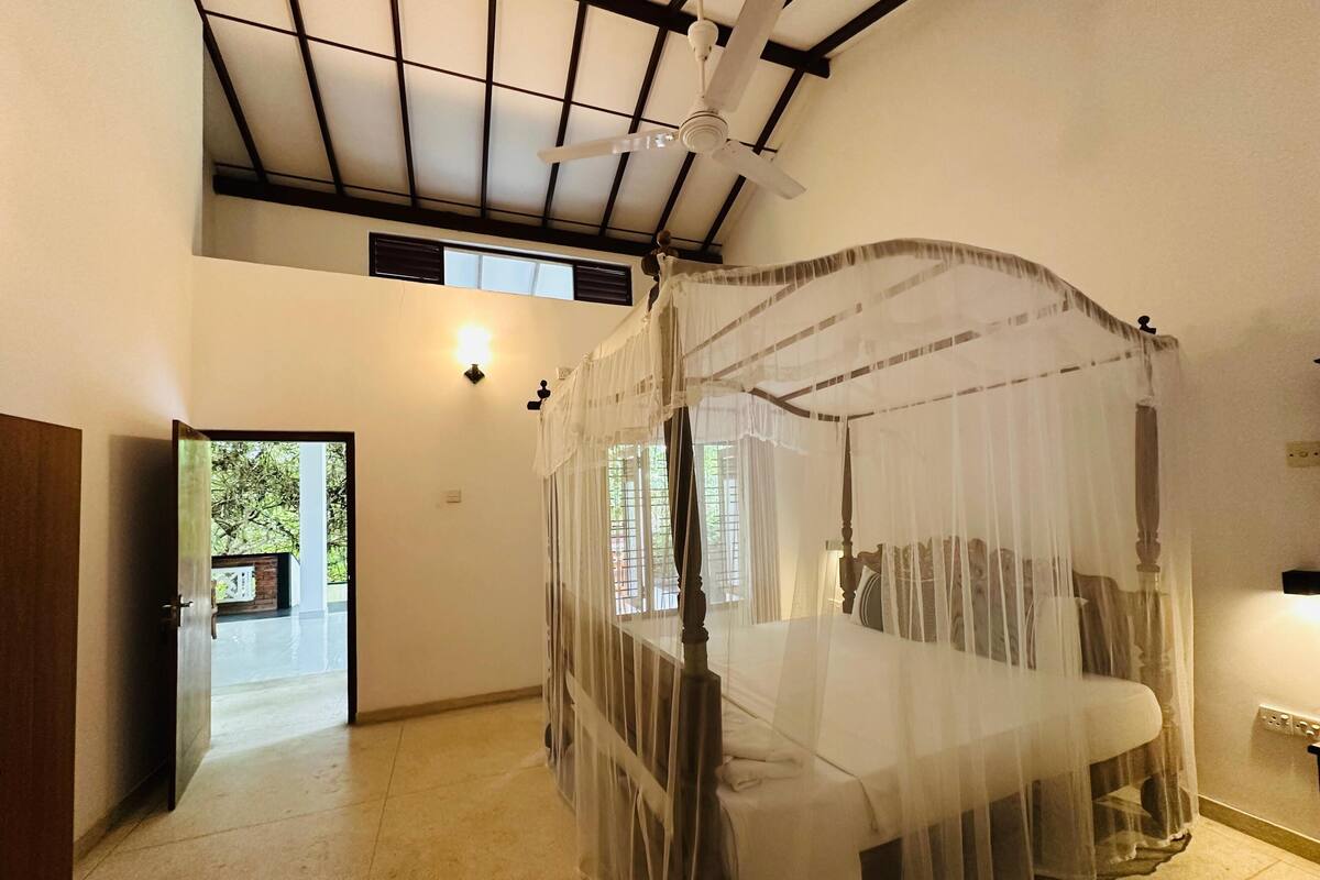 A room with a four-poster bed, a ceiling fan, a wall-mounted heater, and a window with a view of greenery outside.