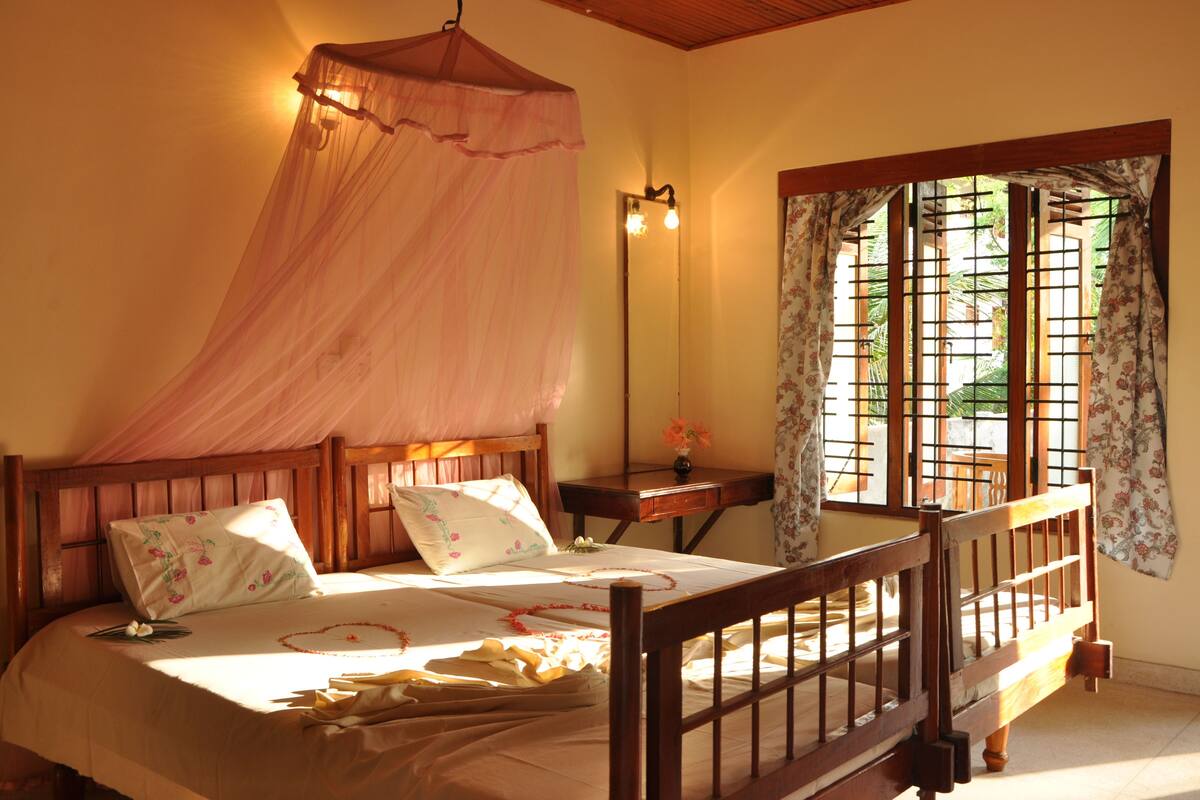 A bedroom with a canopy bed, a bedside table with a lamp, and a window with curtains.