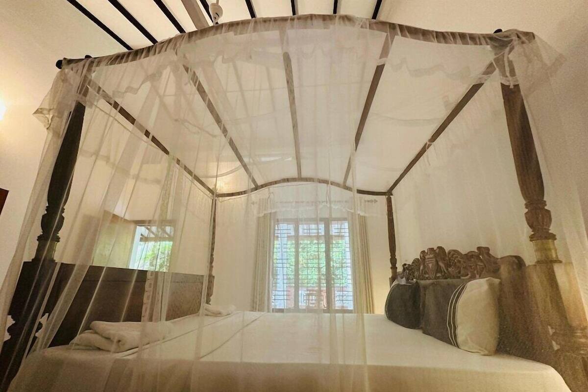 A four-poster canopy bed with a sheer white canopy, wooden headboard, and a view of greenery outside.
