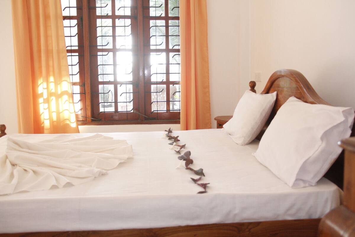 A neatly made bed with white linens and decorative pillows, a wooden headboard, and a window with a view of greenery.