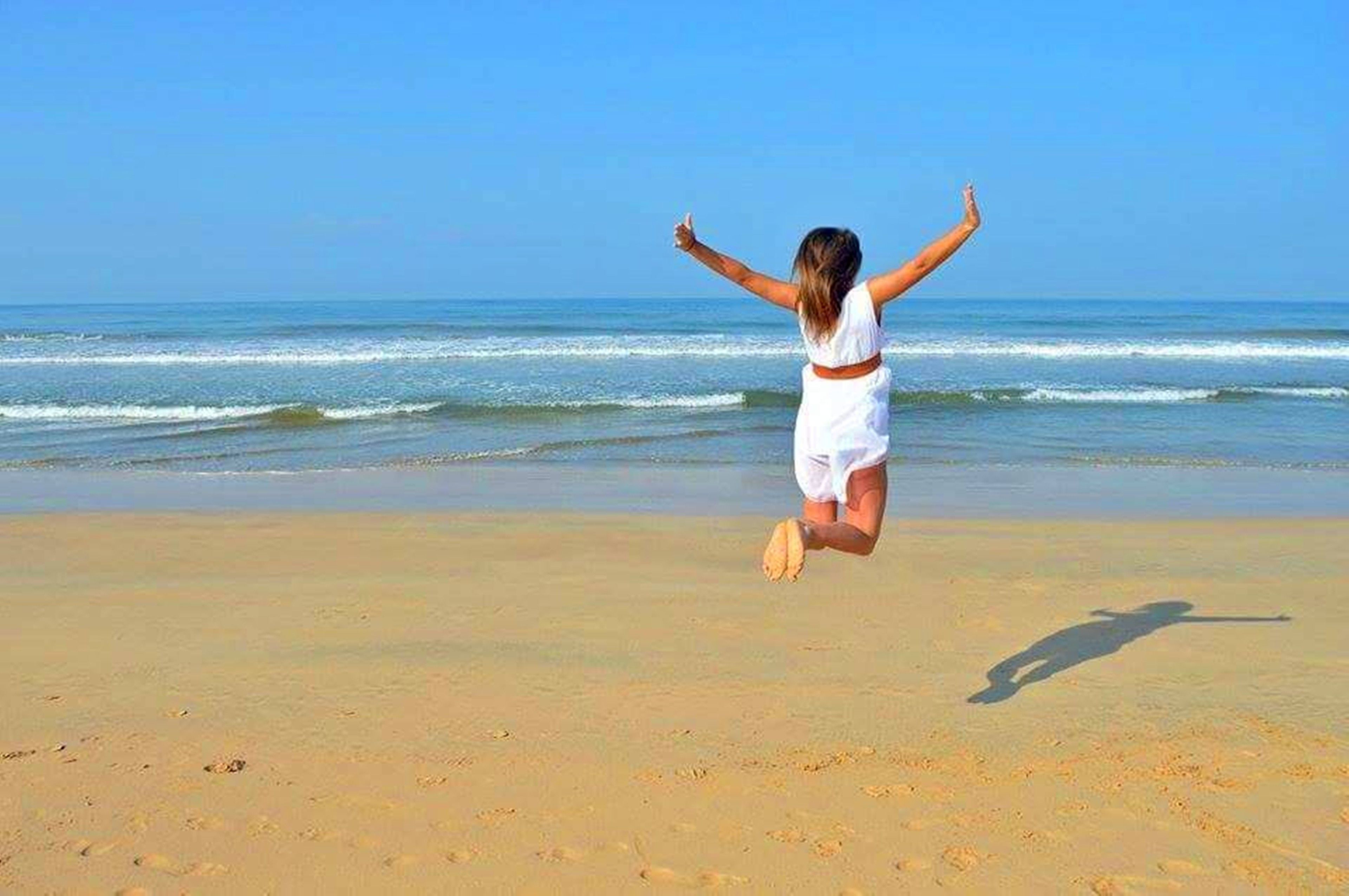 A person jumping on a beach with a clear sky and ocean in the background.