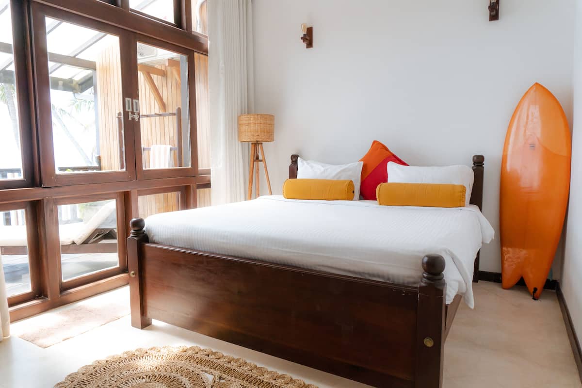 A bedroom with a wooden bed, a surfboard, and a view through large windows.