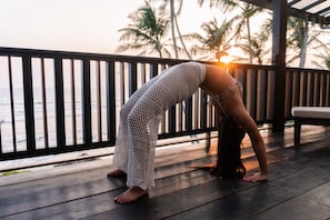 Una persona realiza una postura de yoga en una terraza de madera con vista al océano y palmeras al fondo.