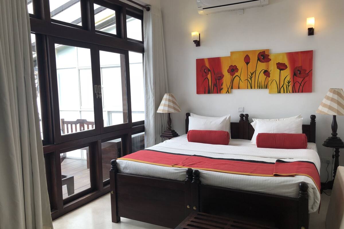 A hotel room with a large window, a bed with red and white bedding, two wall-mounted lamps, and a painting of flowers.