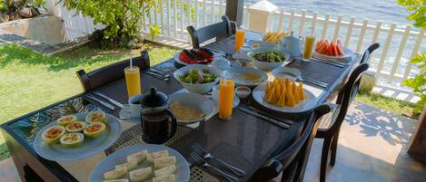A table set for breakfast with various dishes, a view of the ocean, and a white fence in the background.
