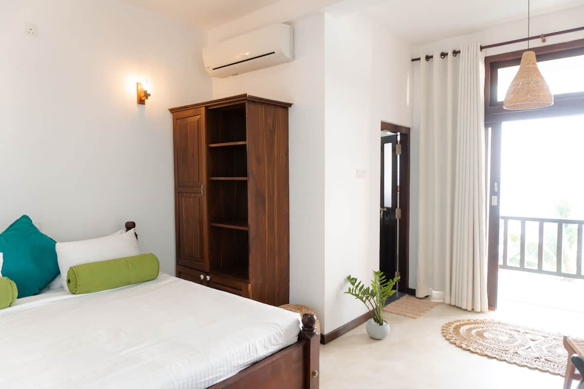 A bedroom with a bed, a wooden wardrobe, a potted plant, and a view of the outdoors through a door.