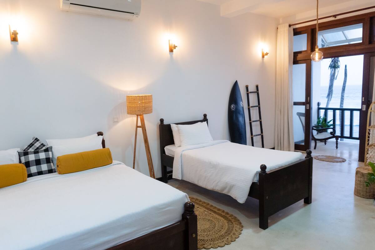 A hotel room with two beds, a surfboard, and a balcony with a view of the ocean.