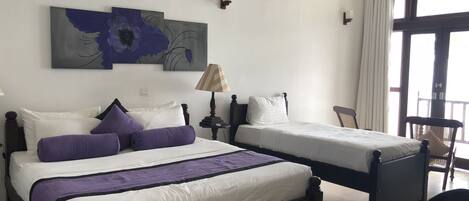 A hotel room with two single beds, a large window with curtains, a wall-mounted air conditioner, and a painting above the beds.