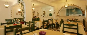 Un comedor con varias mesas y sillas, una cocina visible a través de un arco y plantas colgantes.