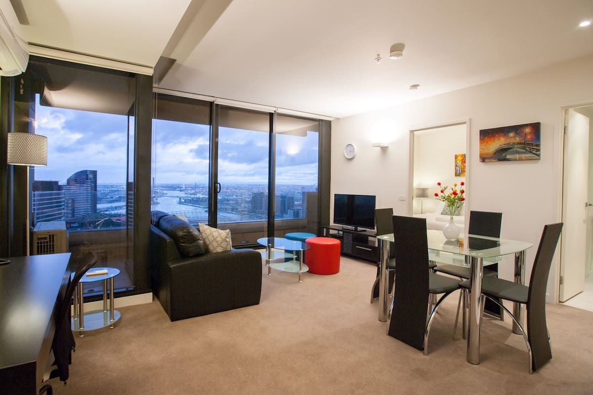 A modern living room with a large window, a sofa, a glass table, and a dining area.