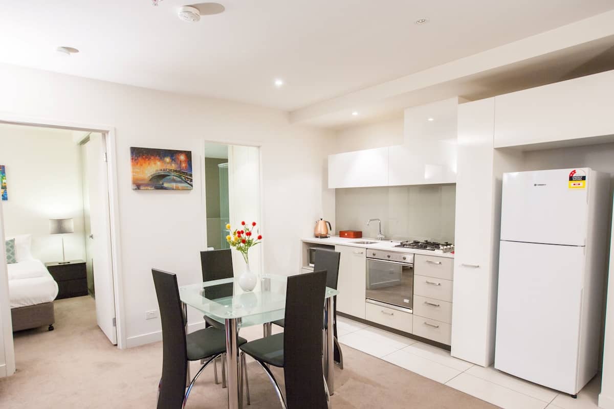 A modern kitchen with white cabinets, a stainless steel refrigerator, and a gas stove. There is a dining area with a glass table and black chairs.