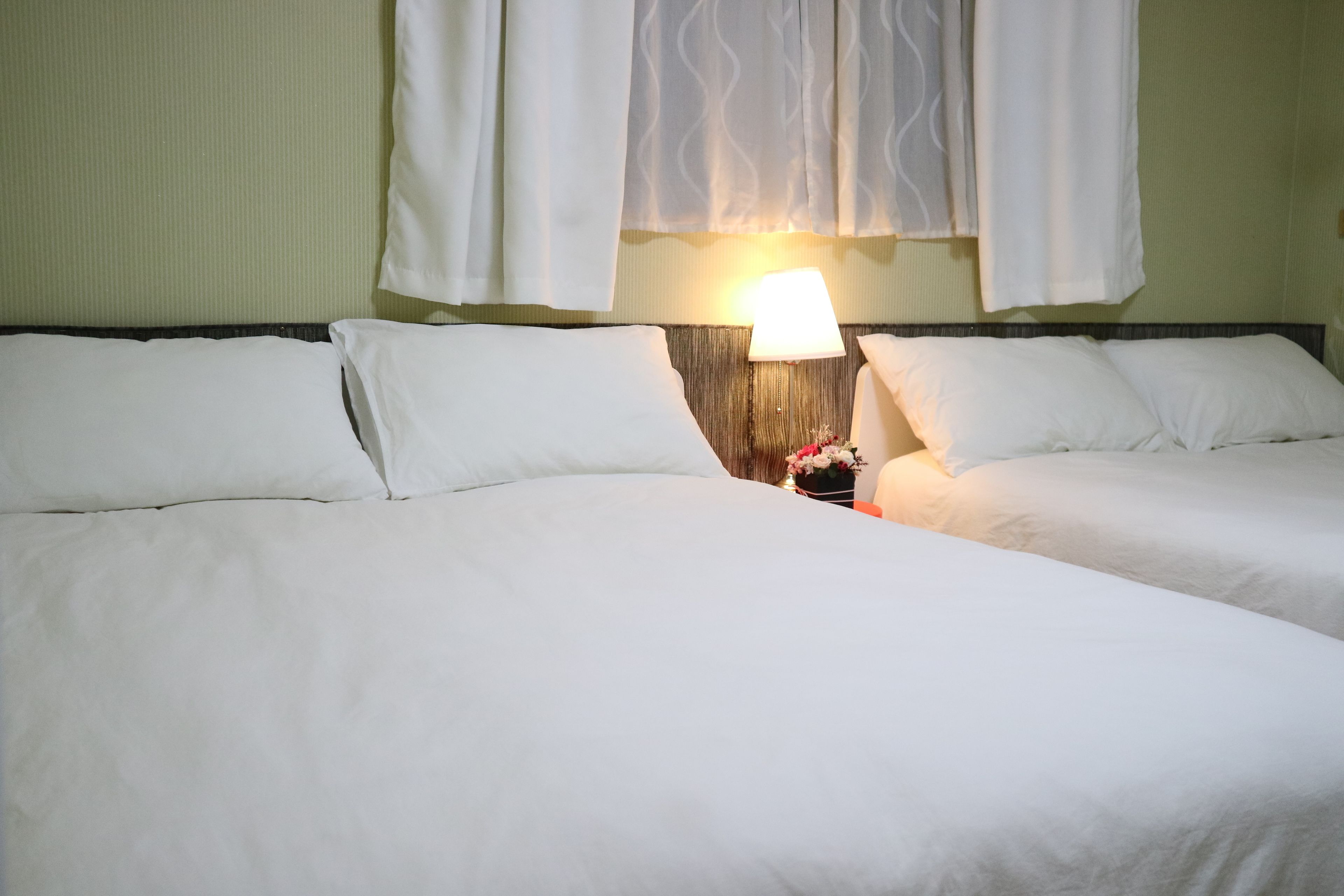 A hotel room with two beds, a nightstand with a lamp, and a window with sheer curtains.