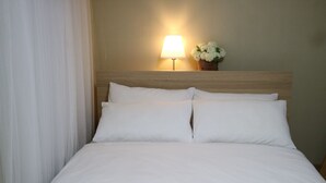Un lit bien fait, avec du linge de lit blanc, une tête de lit en bois, une lampe fixée au mur et un petit vase contenant des fleurs blanches.