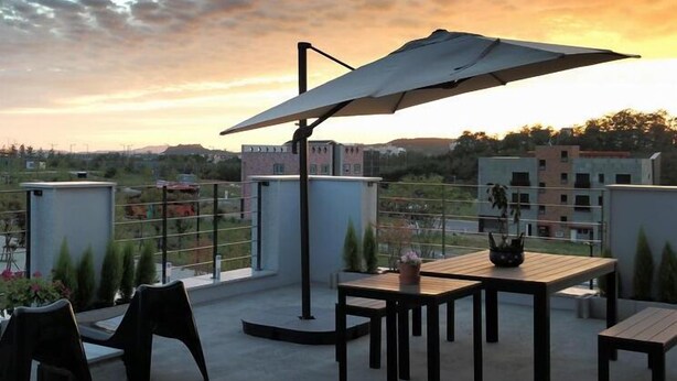 Une terrasse extérieure avec une table et des chaises, un grand parasol, et une vue sur des bâtiments et des collines en arrière-plan.
