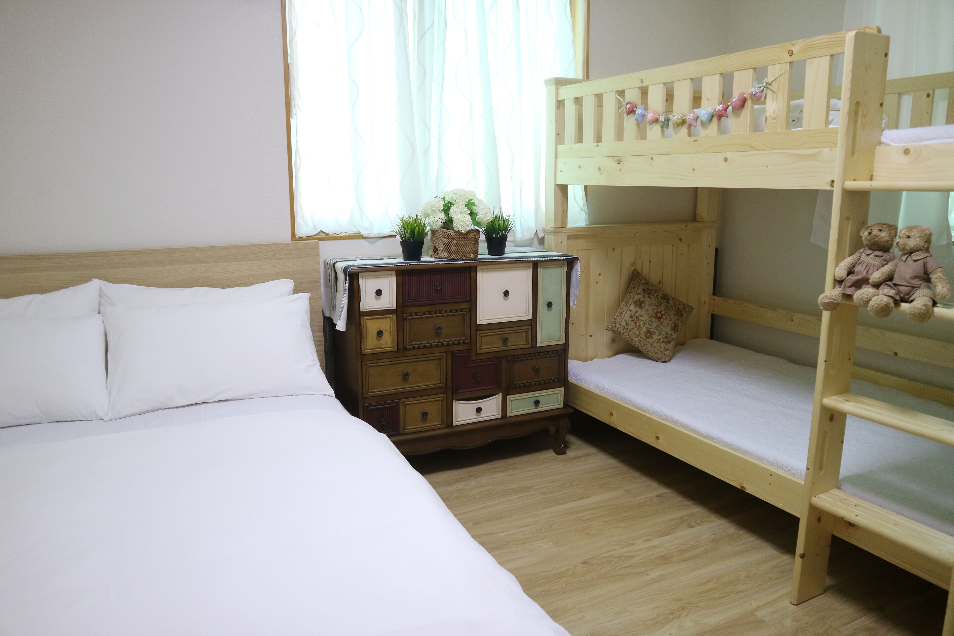Un lit superposé avec une tête de lit en bois, une commode avec de nombreux tiroirs et une fenêtre ornée de rideaux légers.