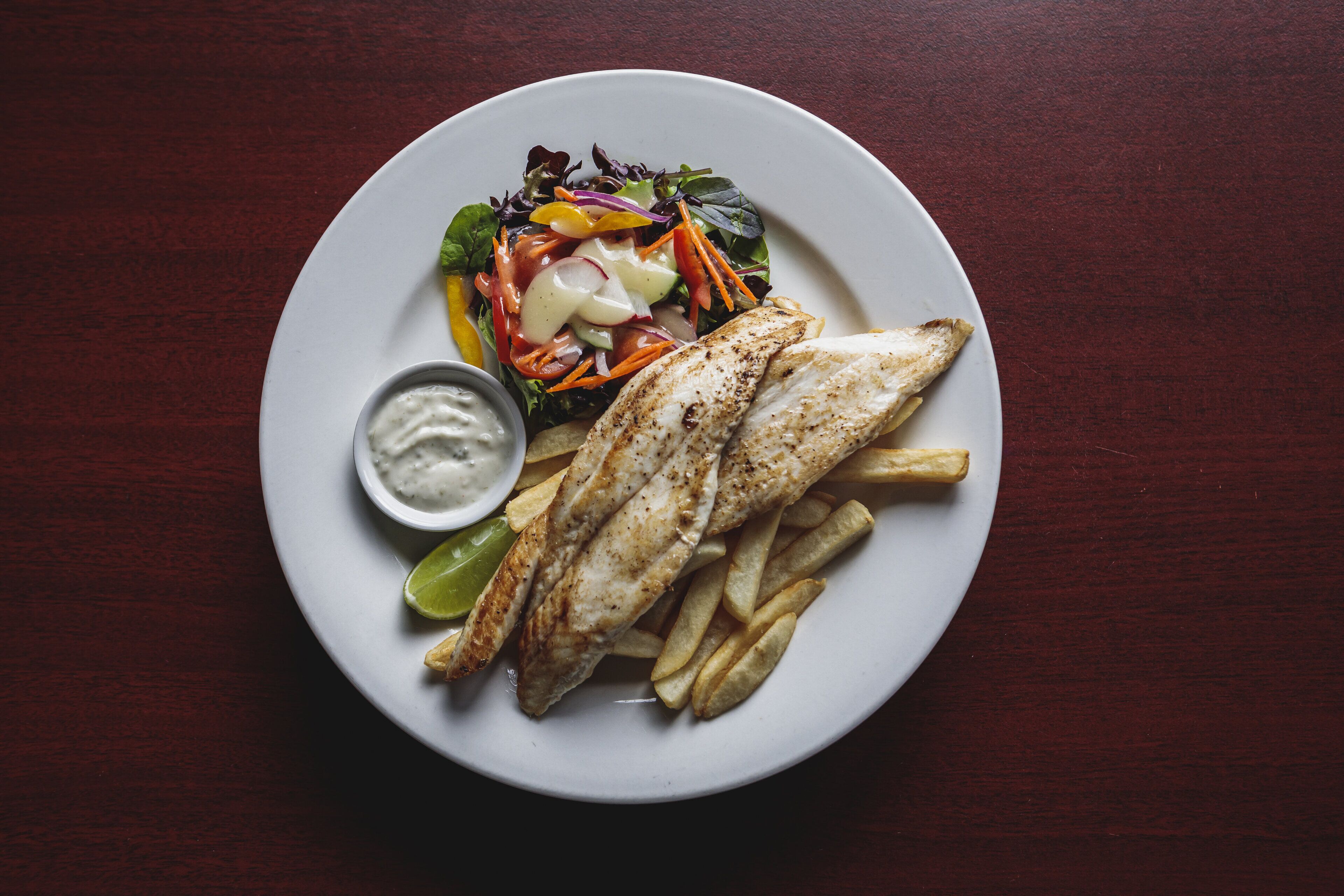 Un plato de pescado a la parrilla, papas fritas y una ensalada con un recipiente pequeño de salsa.