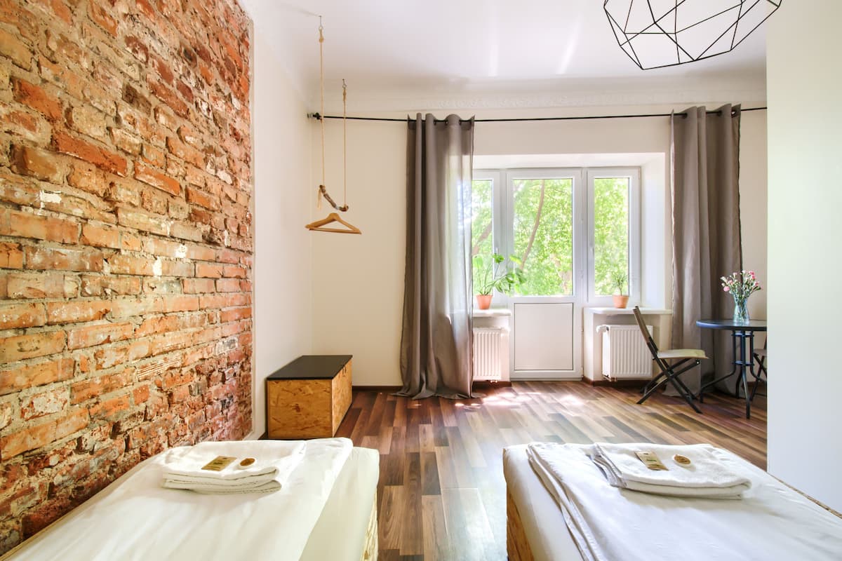 A room with a brick wall, two beds, a hanging chair, a table with a vase, and a window with curtains.
