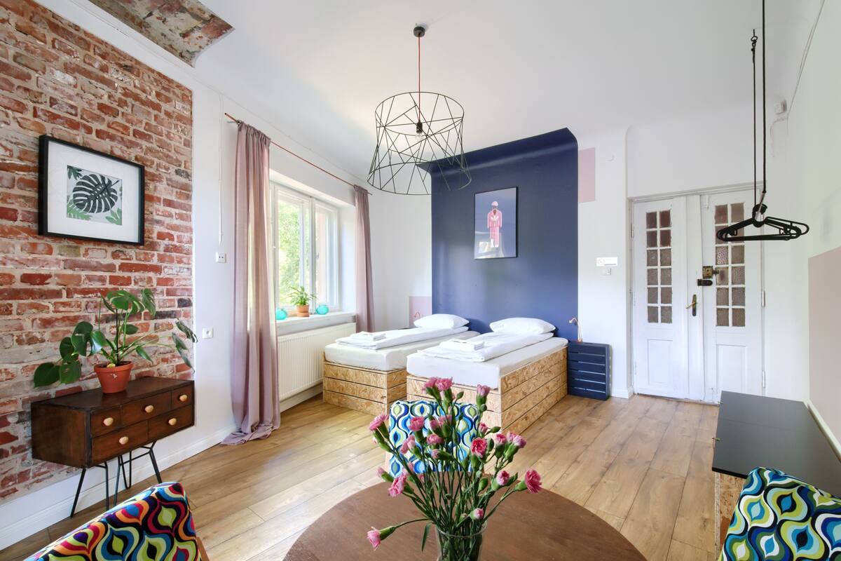 A room with a brick wall, a bed with white bedding, a desk with a plant, and a hanging light fixture.