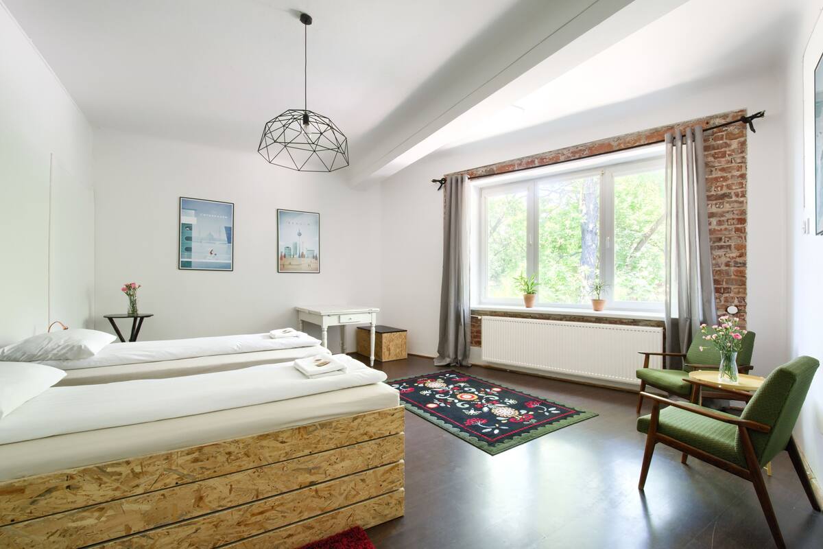 A bedroom with two beds, a desk, a chair, a window with a view of trees, and a hanging light fixture.