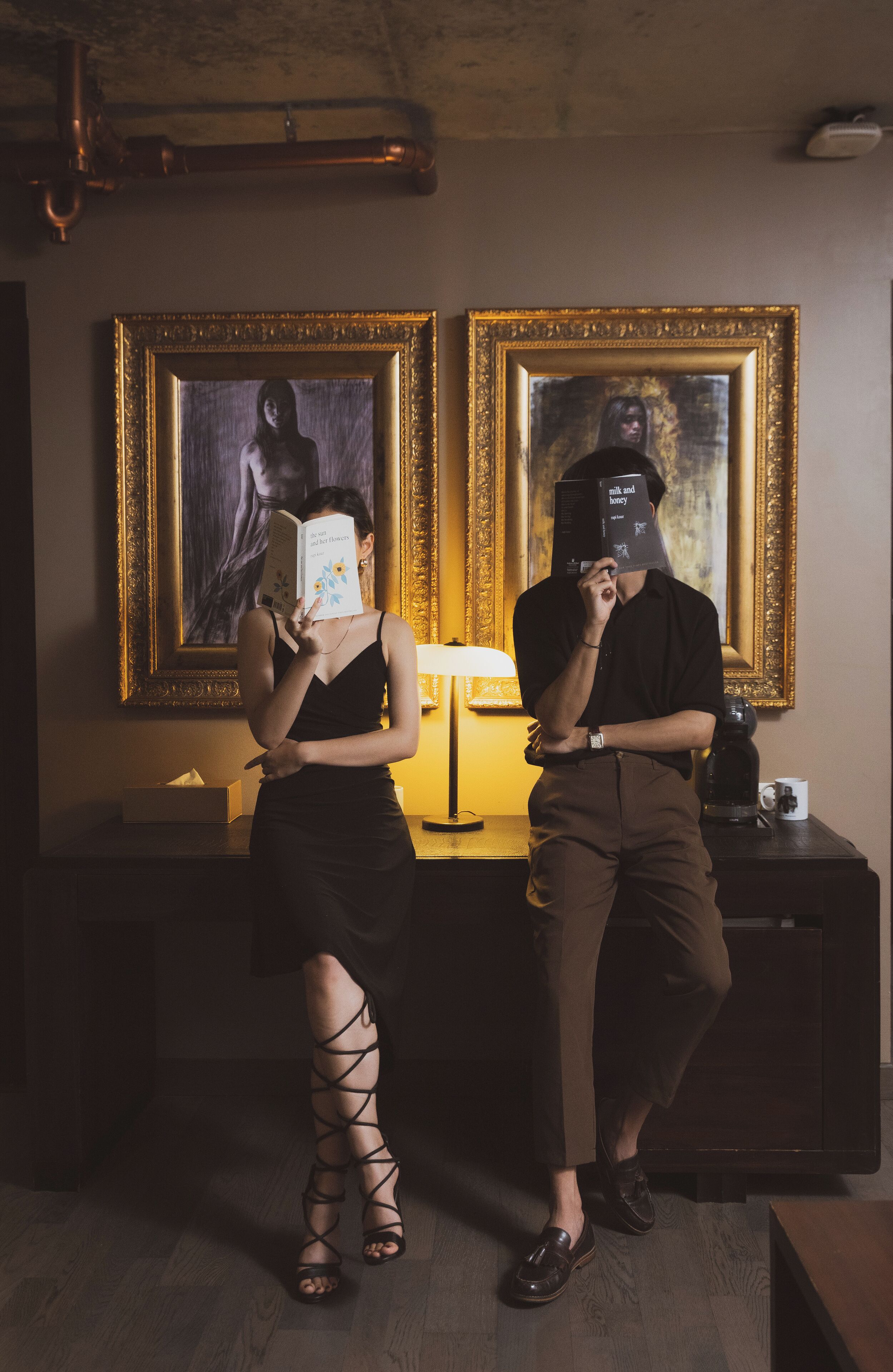 Two people standing in a room with a lamp, a coffee maker, and framed artwork on the wall.