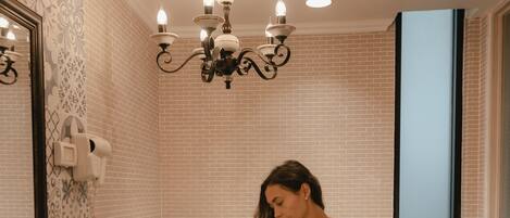 Une femme vêtue d’une robe de couleur claire est assise sur une baignoire dans une salle de bain aux murs décorés de motifs, ornée d’un lustre et d’un miroir.