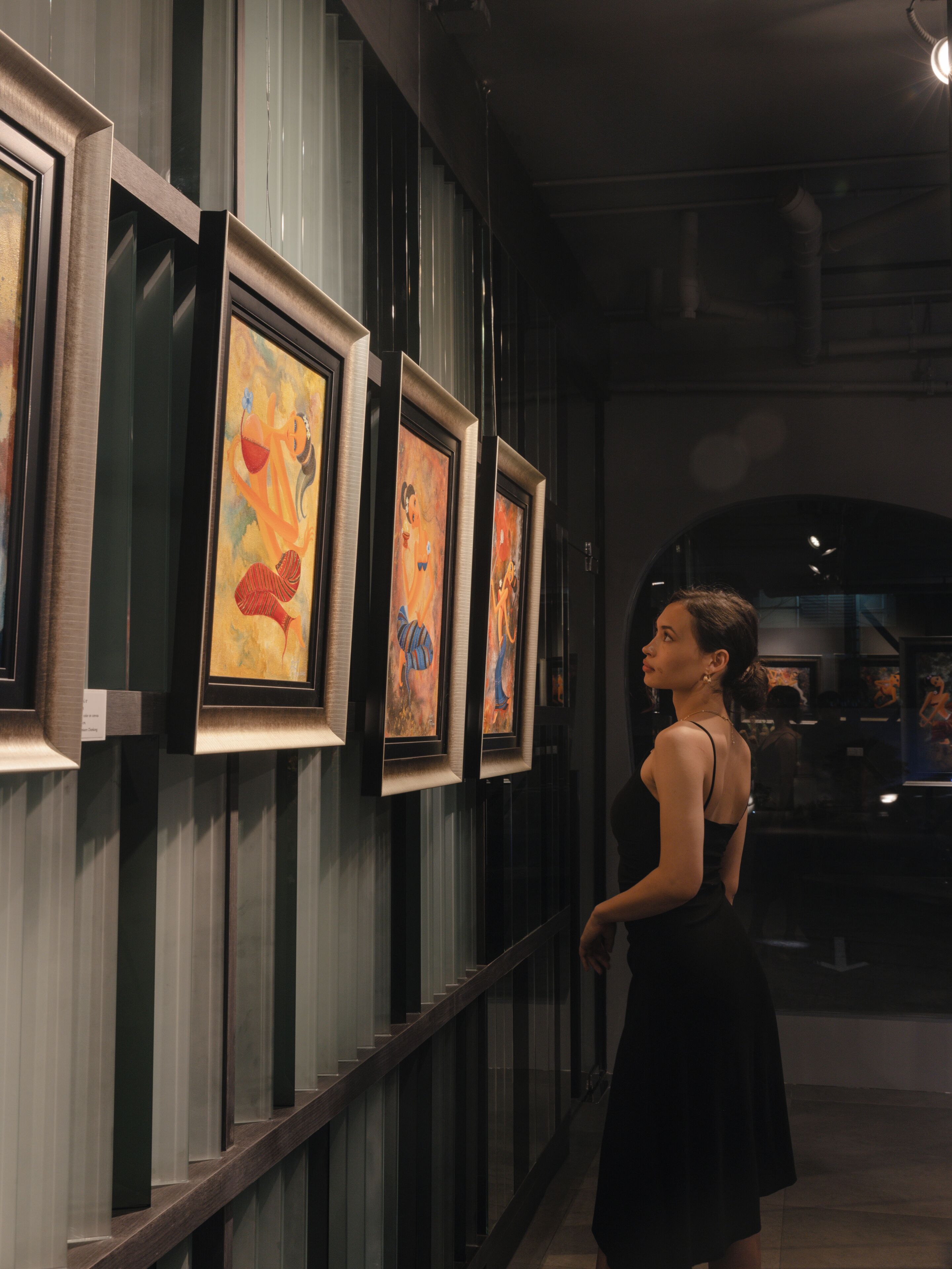 A corridor with framed artworks on the wall, a staircase, and a person in a black dress.