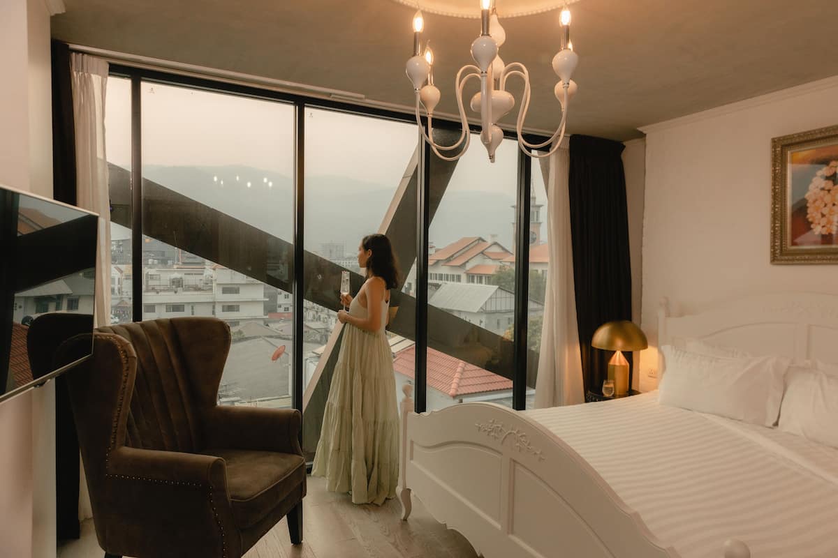 A bedroom with a large window, a bed, a chair, and a nightstand with a lamp.