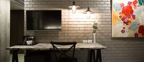 A kitchen with a brick wall, a flat-screen TV, pendant lights, a dining table, and a floral painting.