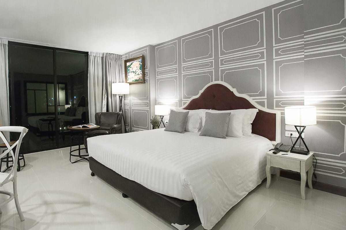 A modern hotel room with a large bed, two bedside tables with lamps, a chair, and a view of the city through a large window.