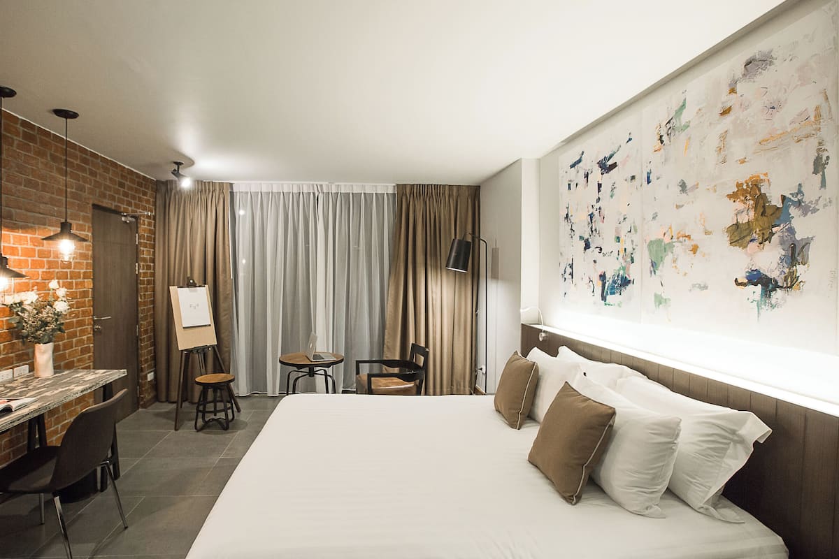 A hotel room with a large bed, a desk with a laptop, a chair, and a small table. There is a brick wall, a window with curtains, and a painting on the wall.