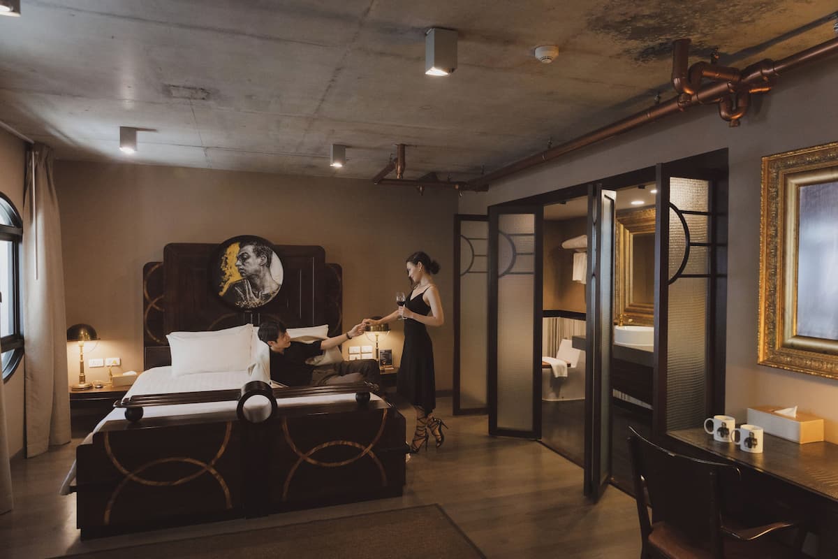 A hotel room with a bed, a desk, a chair, a nightstand, a lamp, a mirror, and a window.