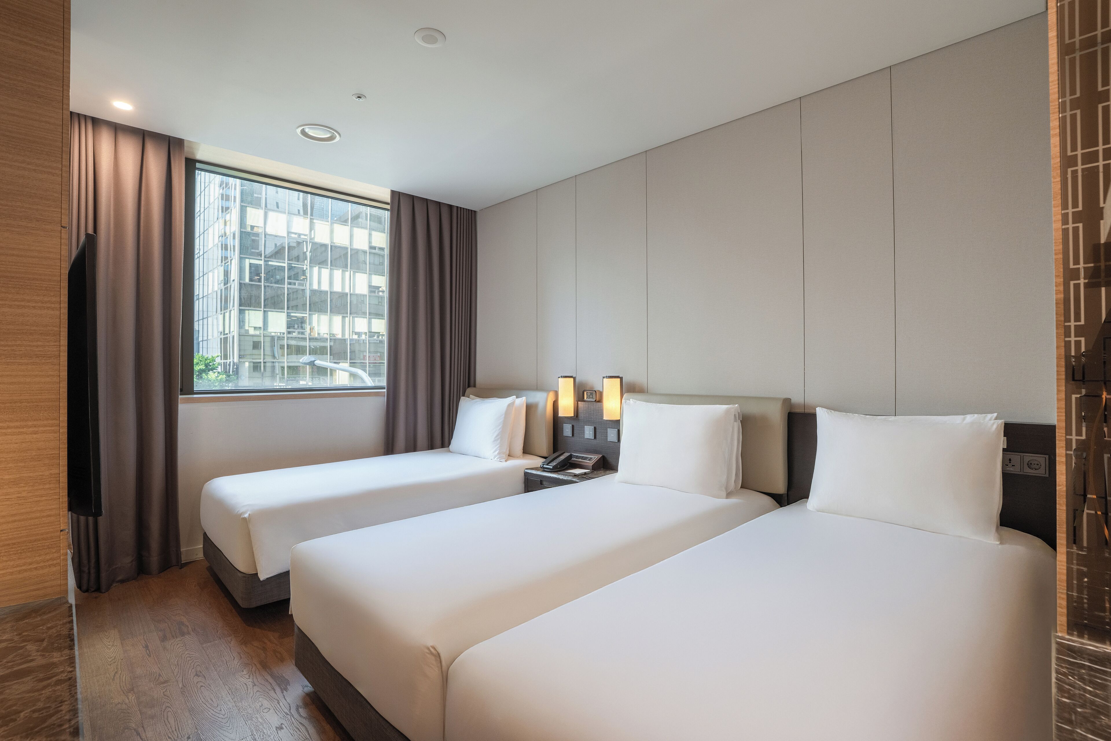 Een hotelkamer met twee bedden, een televisie, een raam met gordijnen en uitzicht op de stad.