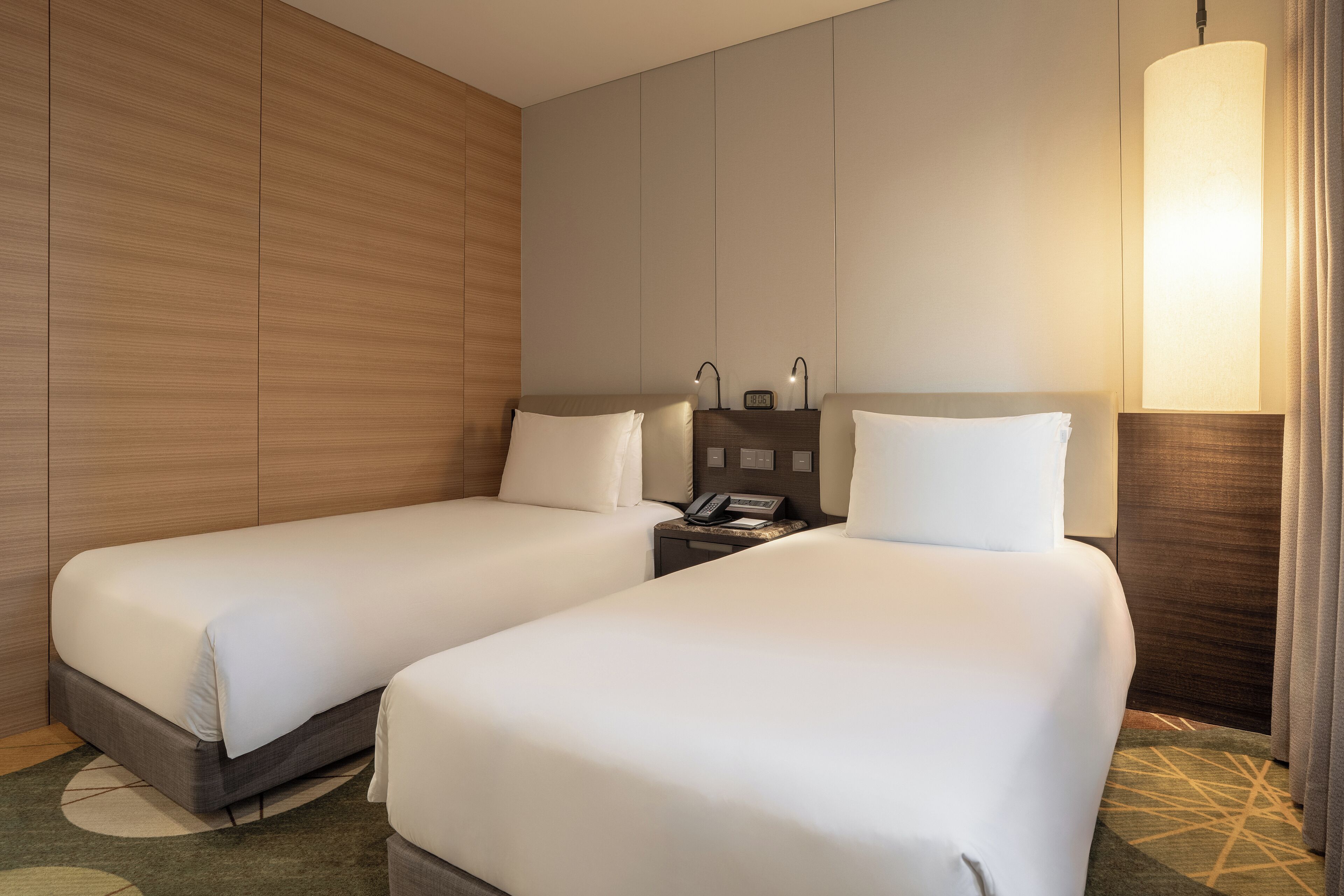 A hotel room with two single beds, a bedside table with a lamp, and a wall-mounted phone.