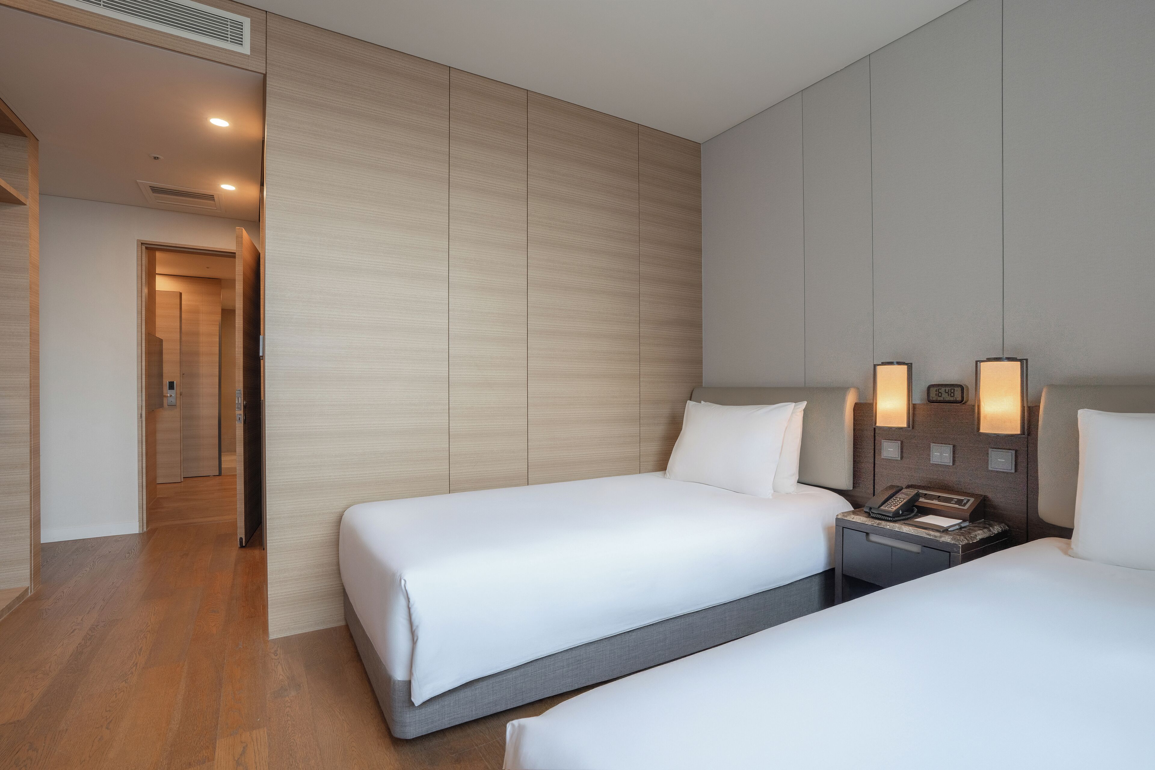 A hotel room with two beds, a wooden floor, a wall-mounted phone, and a nightstand.