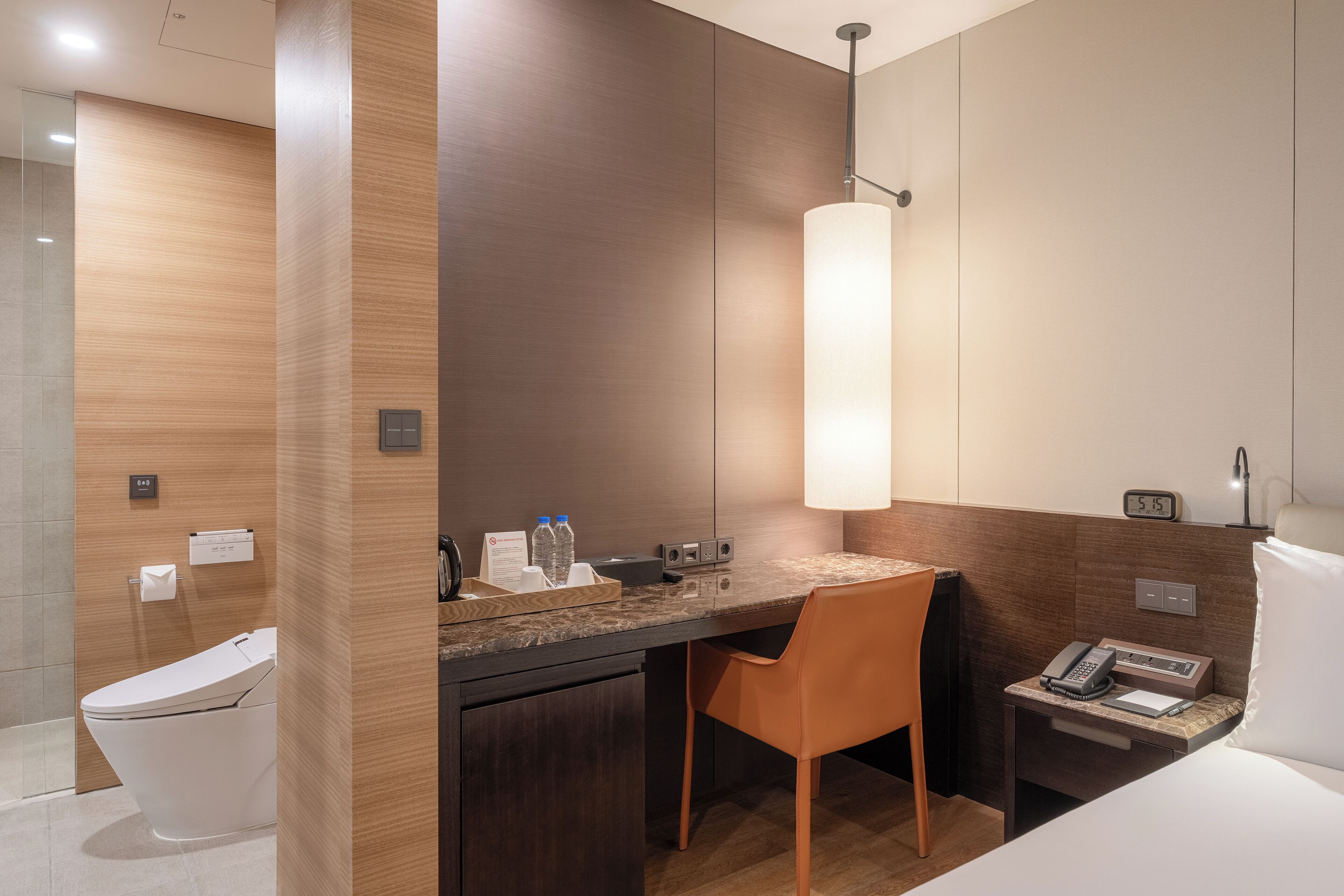Une chambre d’hôtel moderne avec un mur d’accent en bois, un comptoir en marbre, une seule chaise orange, une table de chevet avec un téléphone et une lampe, et des toilettes intégrées.