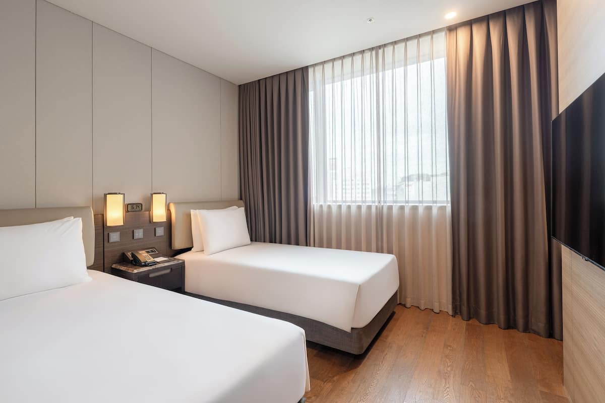 A hotel room with two beds, a large window with curtains, a flat-screen TV, and a bedside table with a lamp and phone.
