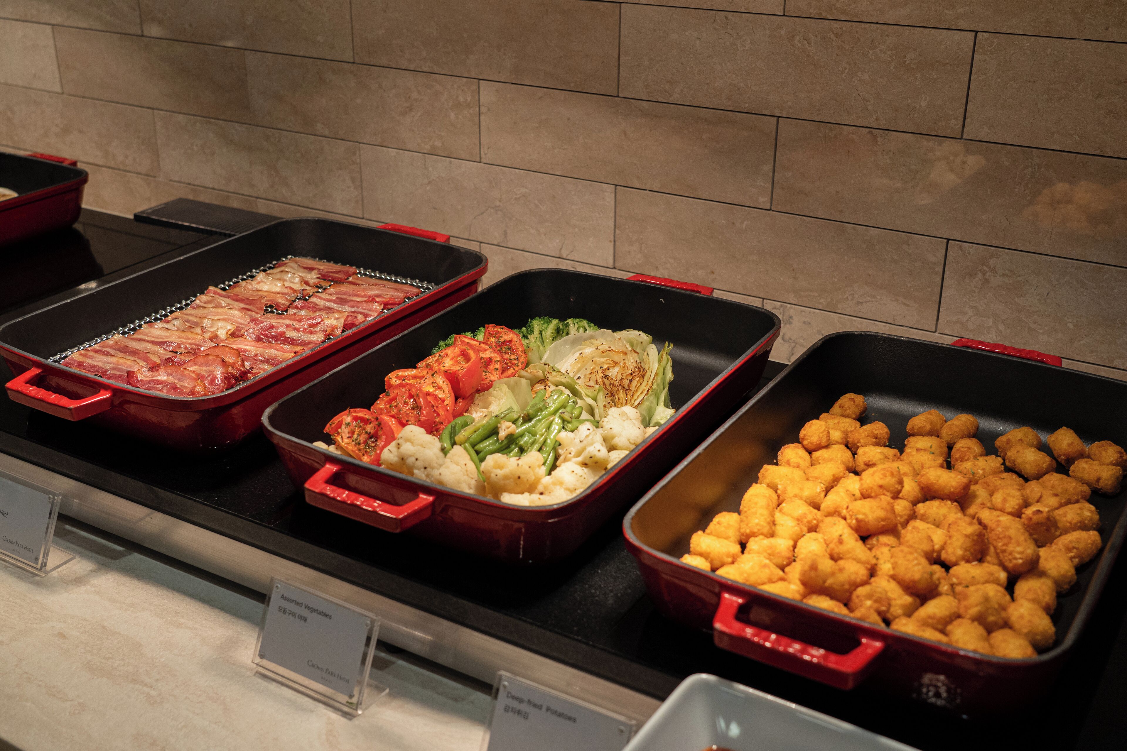 A buffet with various dishes including bacon, vegetables, and tater tots.