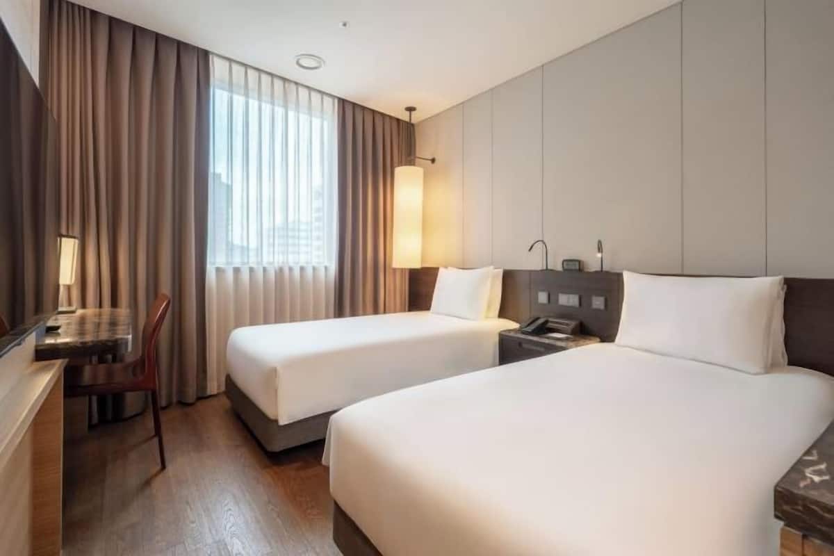 A hotel room with two beds, a desk, a chair, a lamp, and a large window with curtains.
