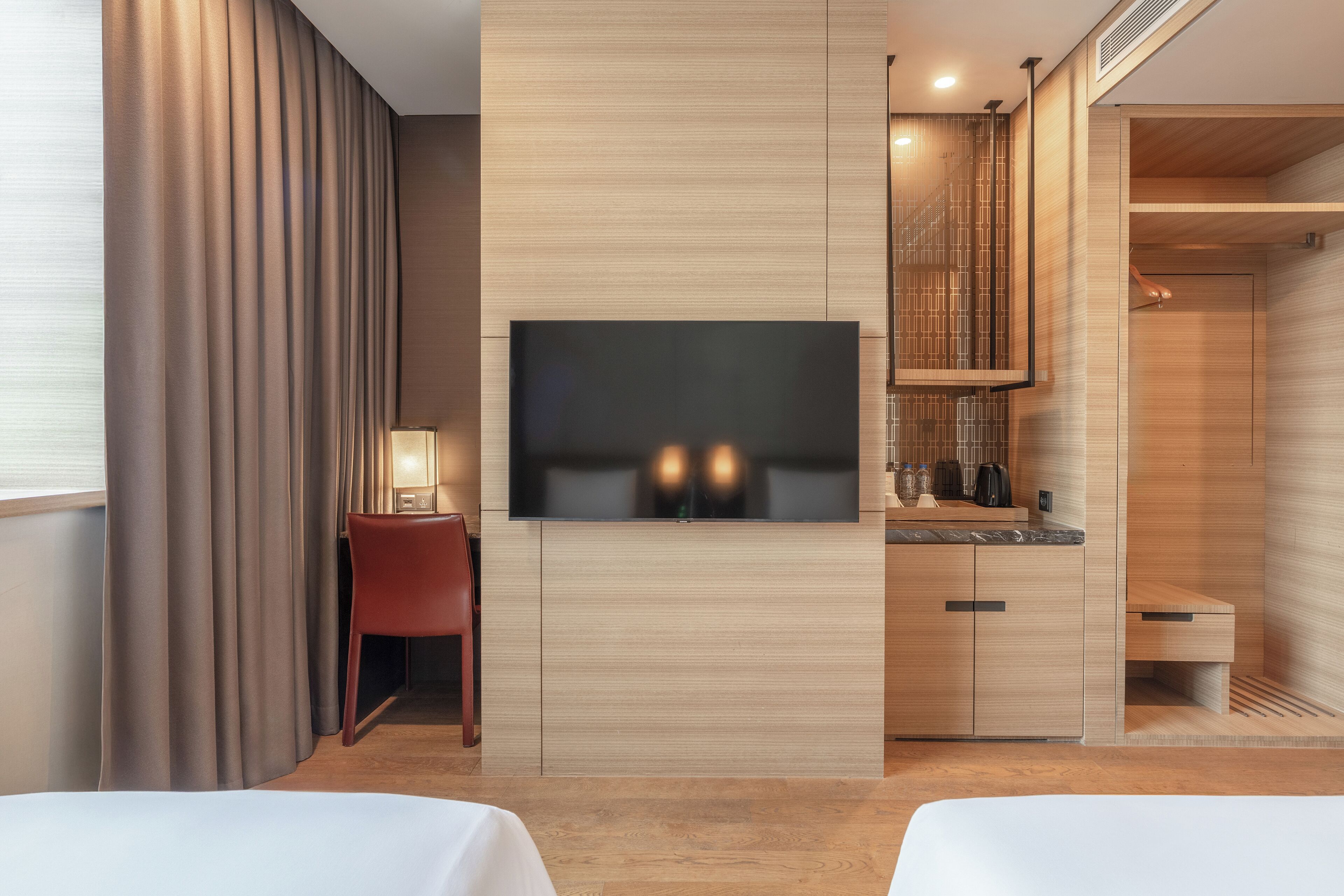 Una habitación de hotel moderna con una televisión de pantalla plana montada en la pared, un armario de madera, un escritorio con una silla y una zona de ducha.