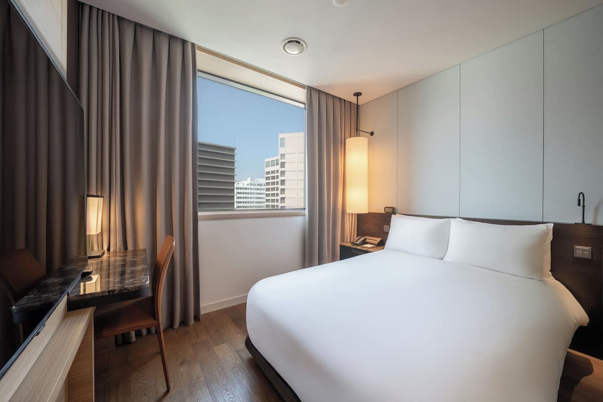 A hotel room with a large bed, a desk with a chair, a lamp, and a view of buildings through the window.