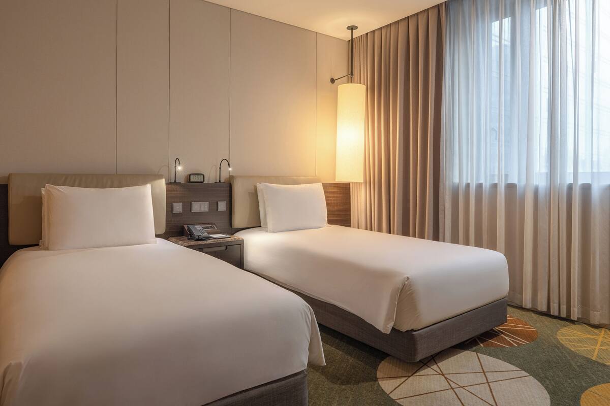 A hotel room with two beds, a nightstand with a lamp, and a view of the outside through sheer curtains.