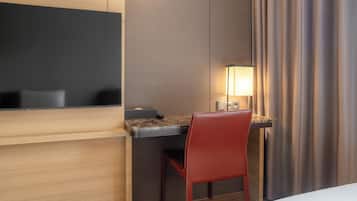 A hotel room with a desk, a chair, a TV mounted on the wall, and a lamp on the desk.