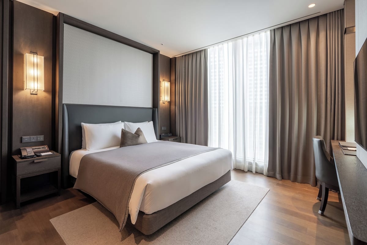 A modern hotel room with a large bed, bedside tables, a desk, and a large window with curtains.