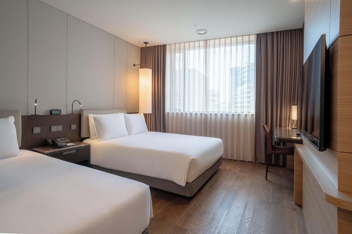 A hotel room with two beds, a desk, a chair, a TV, and a large window with curtains.