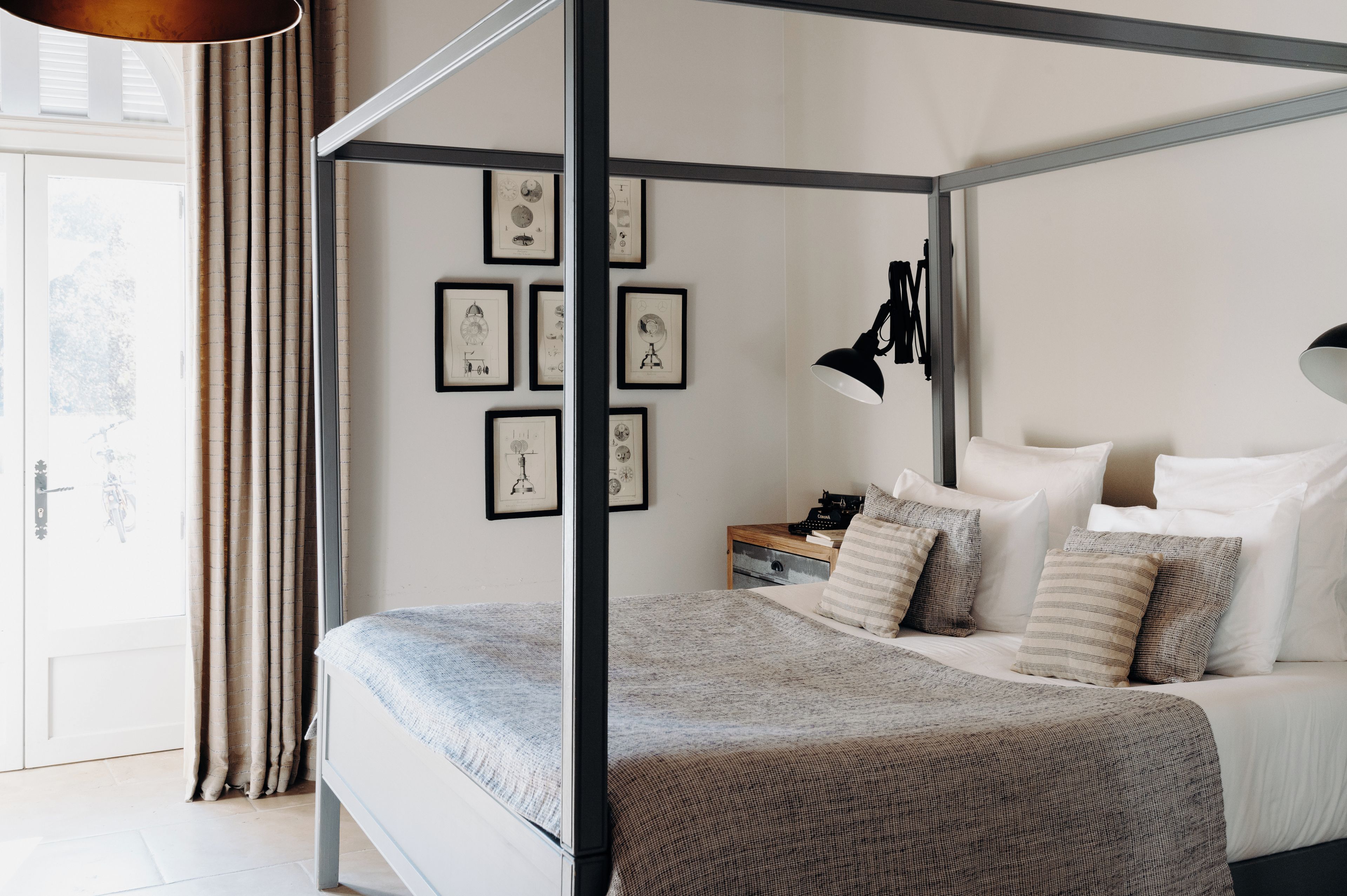 Een hemelbed met een grijsbruin geruit beddegoed, een nachtkastje met een lamp, en lijstwerken aan de wand.