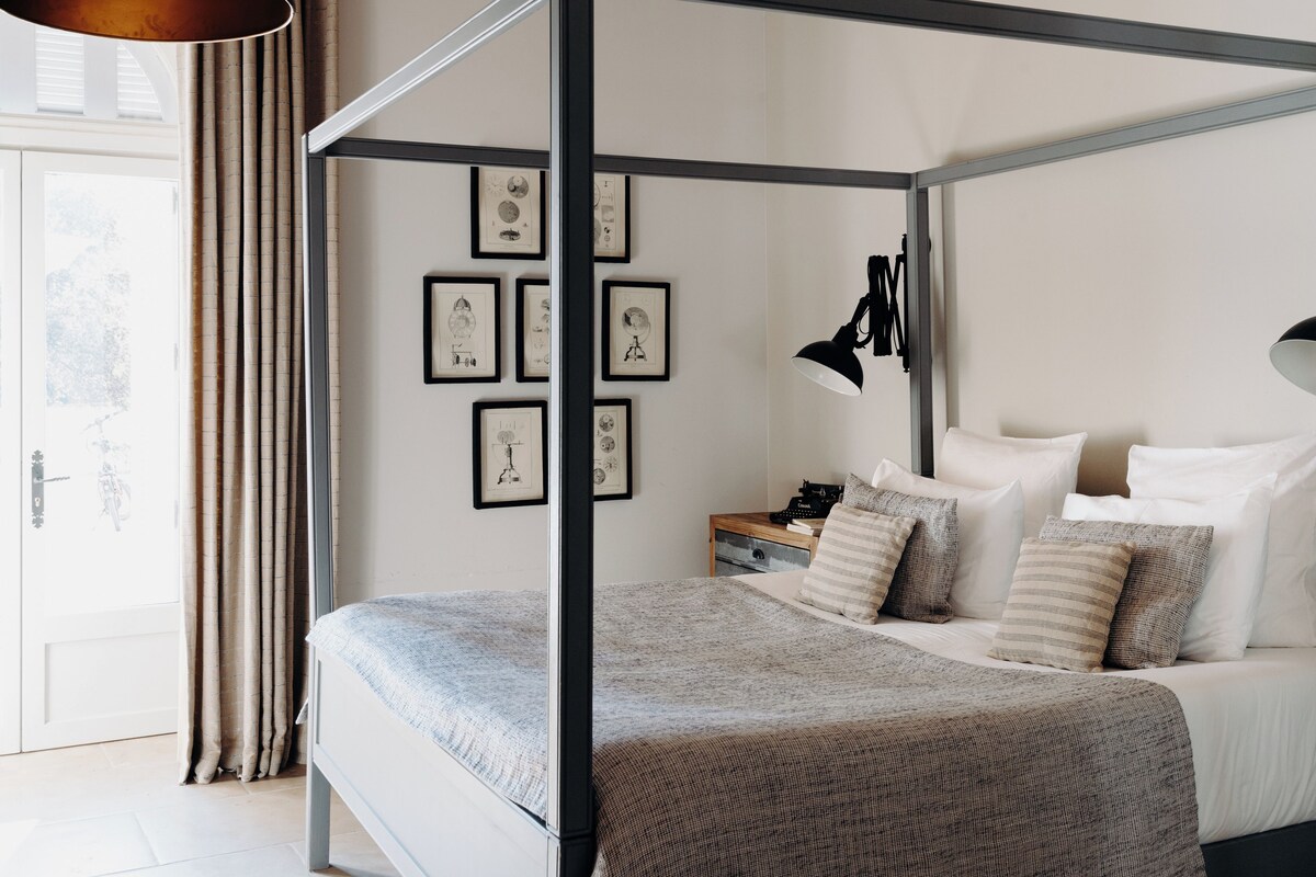 Una cama con dosel con un edredón a cuadros en tonos grises y marrones, una mesita de noche con una lámpara, y cuadros en la pared.