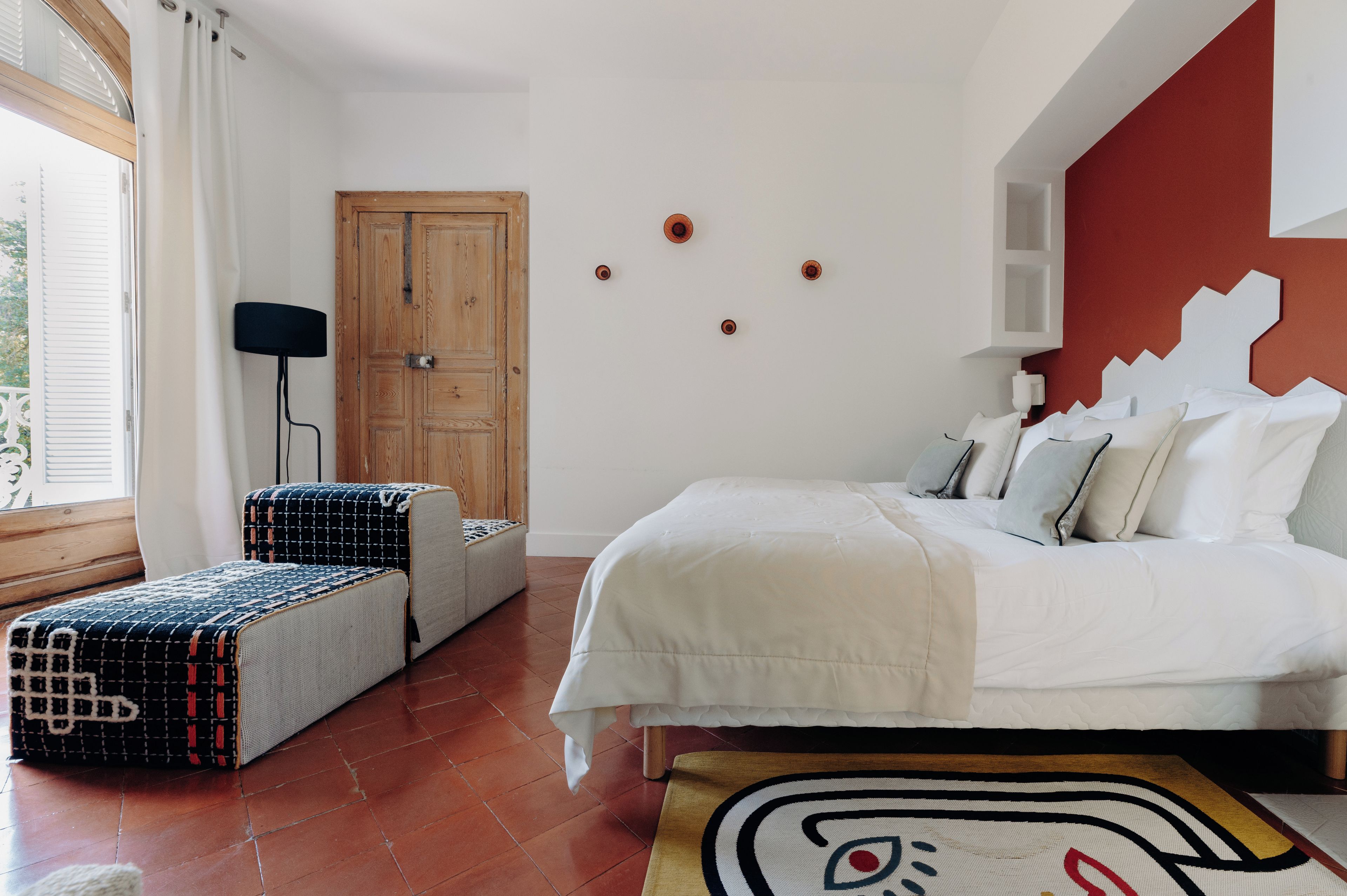 A bedroom with a bed, a wooden door, a window with curtains, and a wall-mounted light fixture.